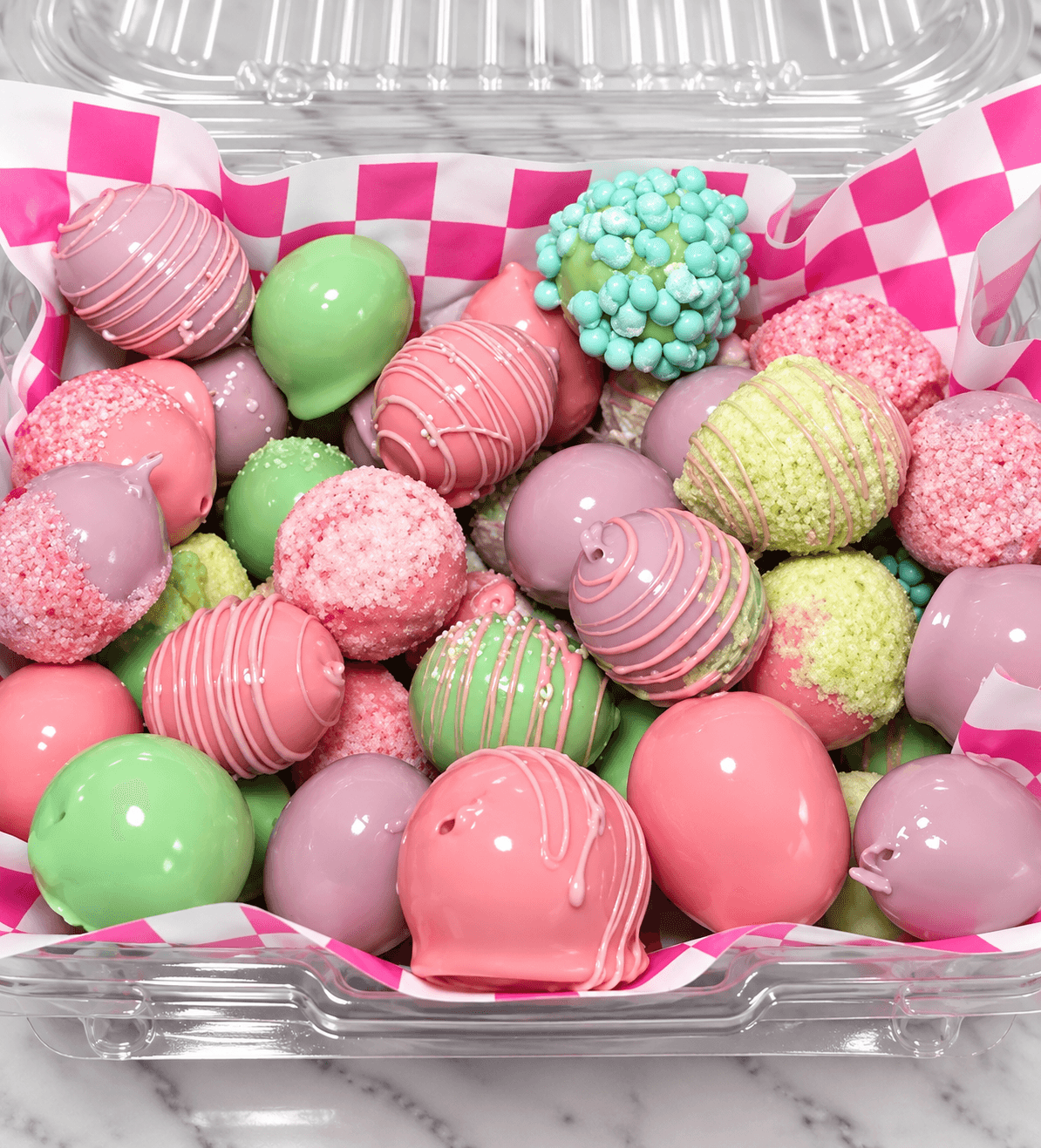Assorted pastel cake pops with drizzles, sprinkles, and smooth glazes in a clear plastic container.